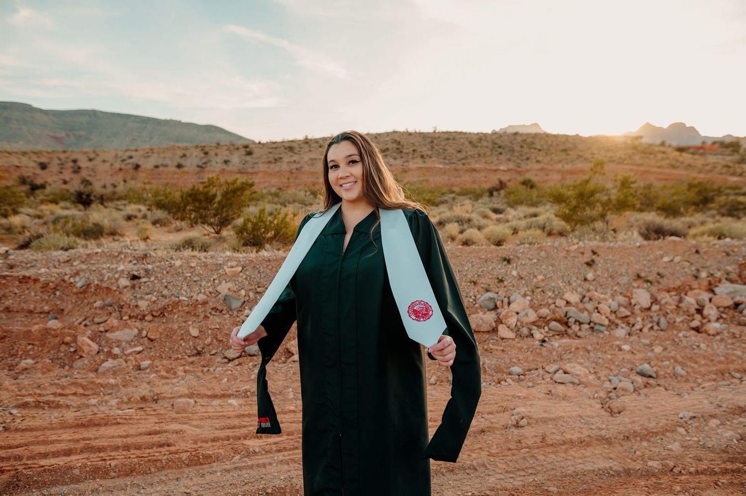 Graduate in black cap and gown poses at sunset in desert with mountains behind her.