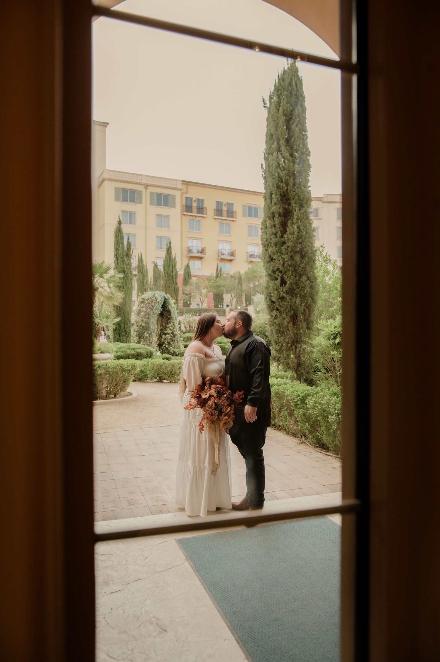 Bride and groom share a tender kiss framed by a window with cypress trees in background.