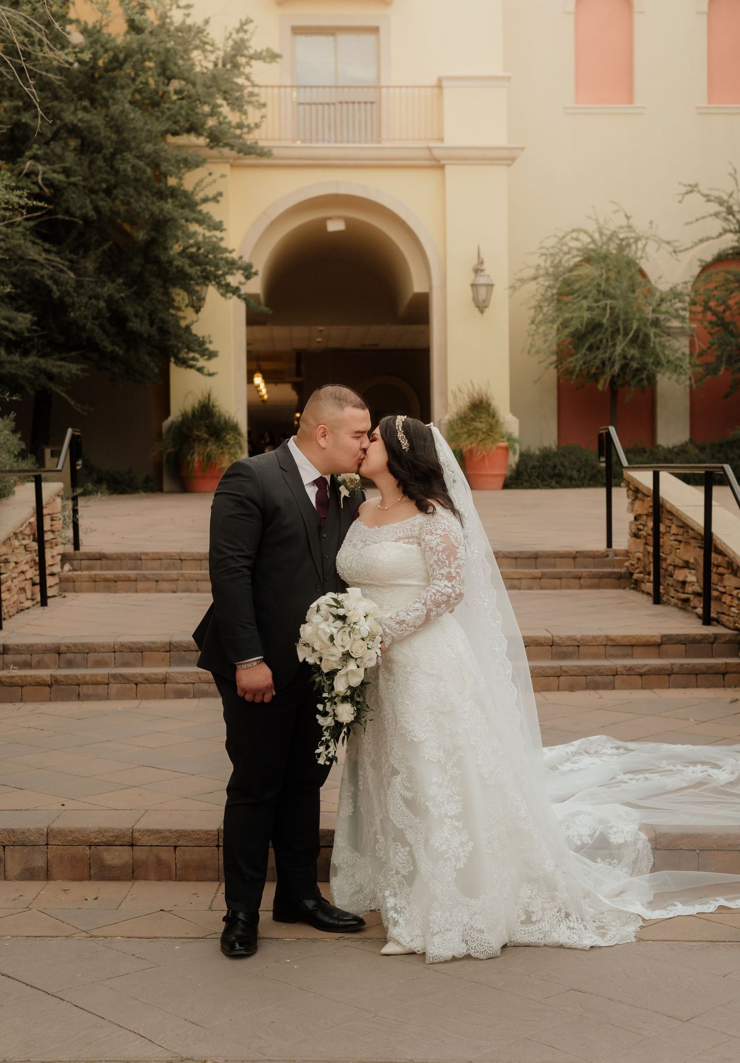 Bride and groom sharing a kiss on steps outside a Spanish-style building with bouquet.