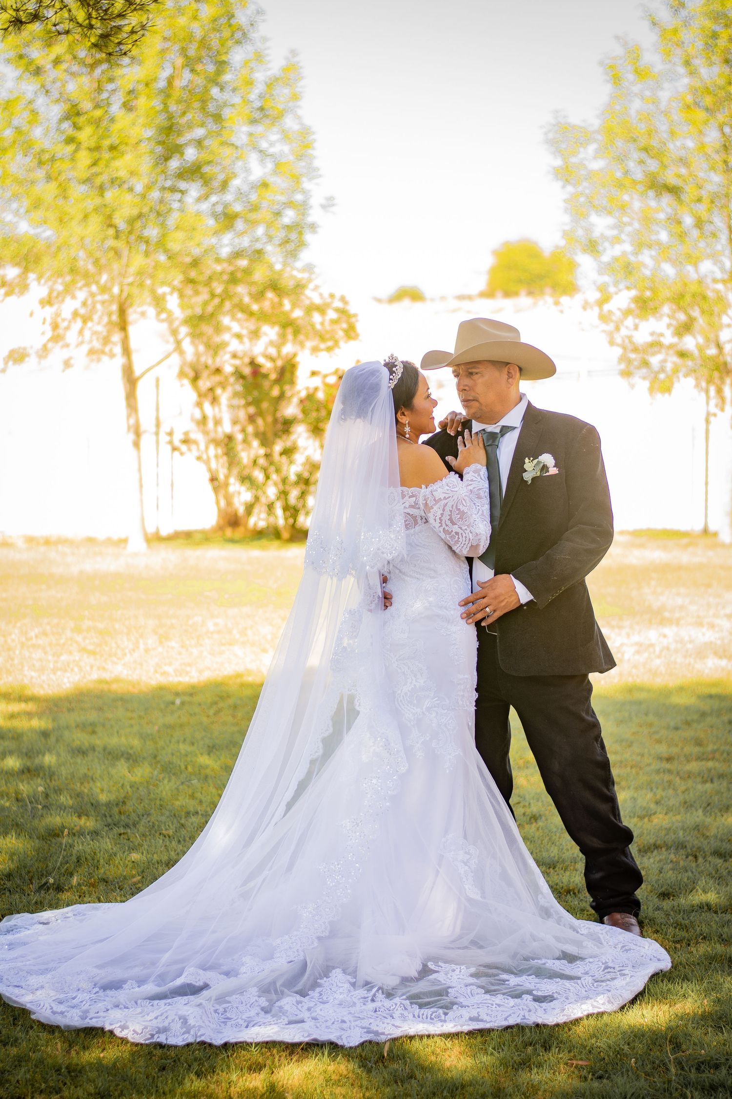 Bride in lace gown with long veil and groom in cowboy hat share a tender moment at golden hour.