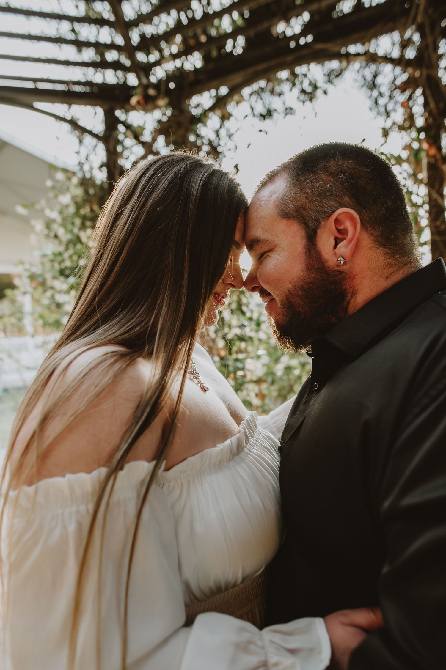 Bride and groom touch foreheads intimately under a pergola in warm golden light with a soft bokeh background.