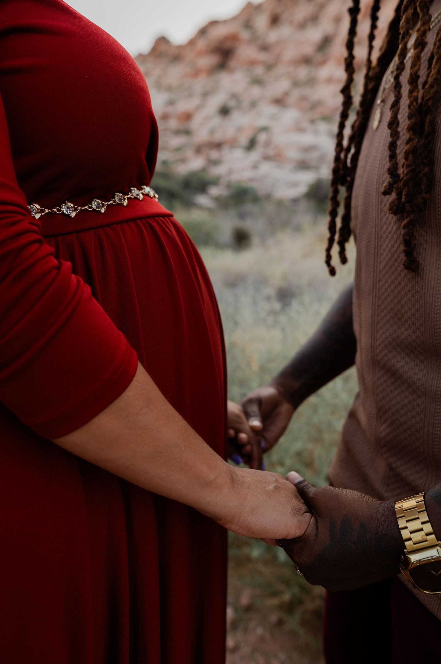 Pregnant woman in red maternity gown holds hands with partner in desert setting.