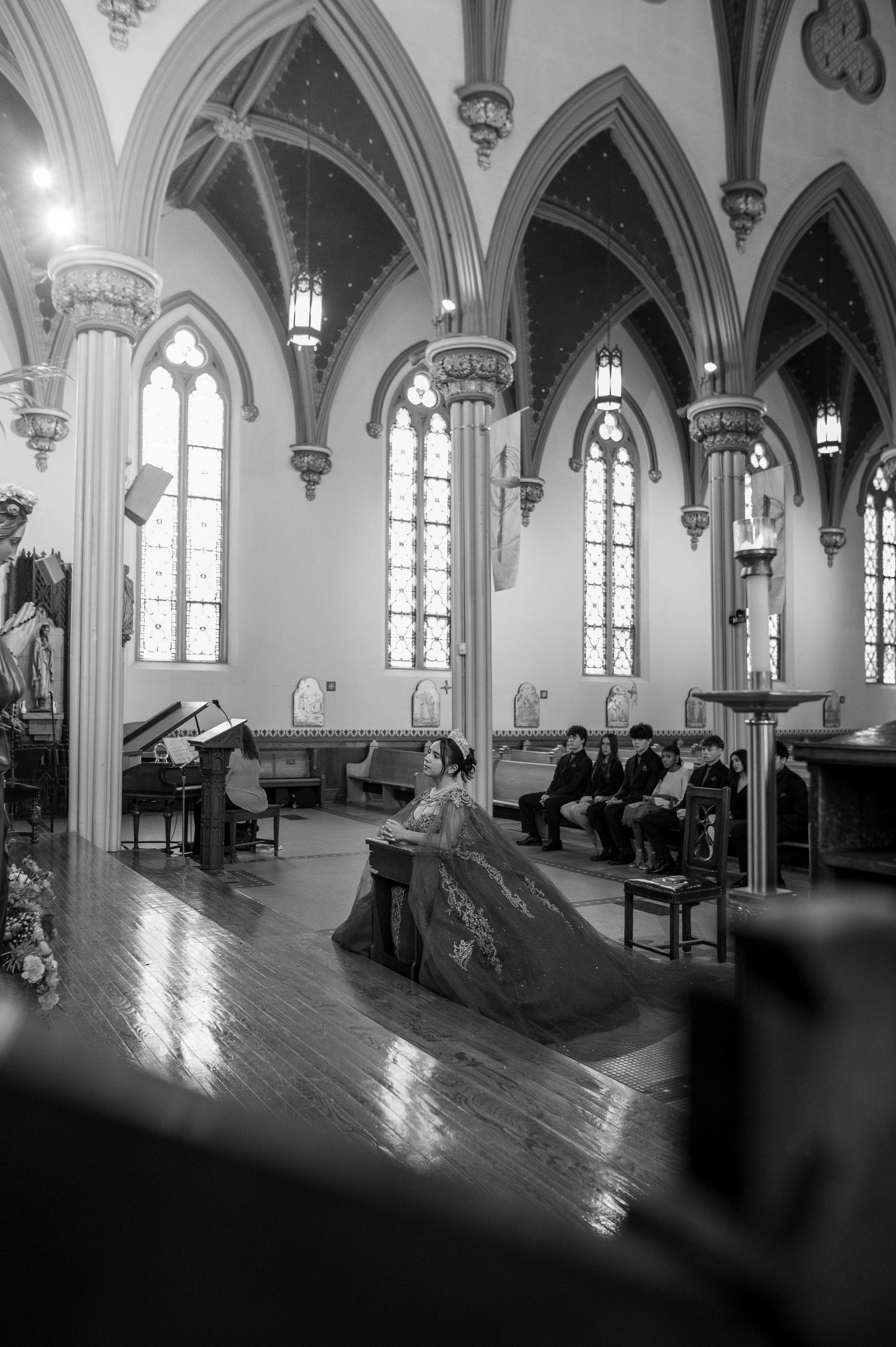 Black and white photo of a quinceañera in a red gown kneeling in a Gothic church aisle.