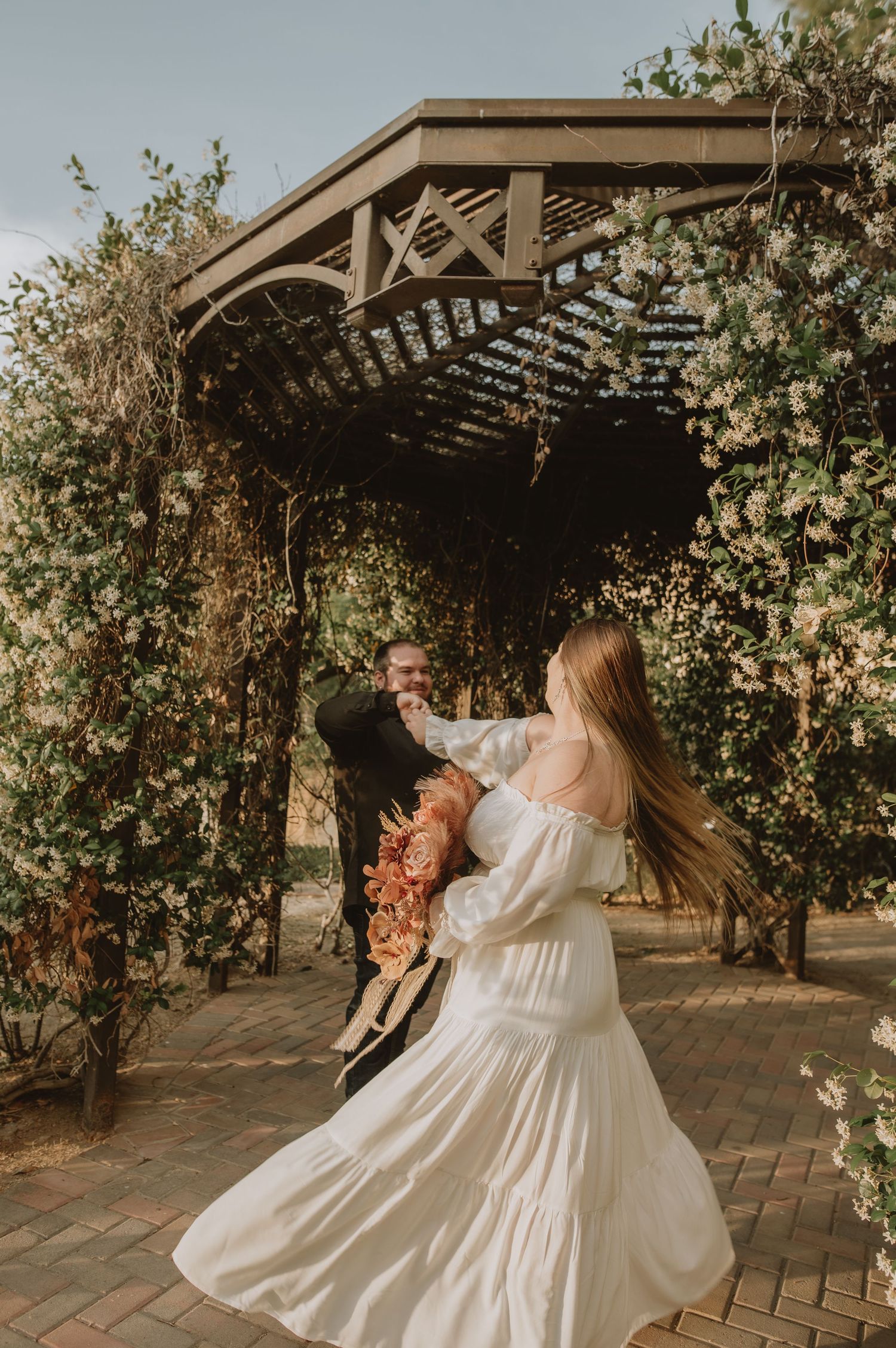 Bride in white off-shoulder gown twirls under floral pergola as groom smiles during outdoor wedding photos.