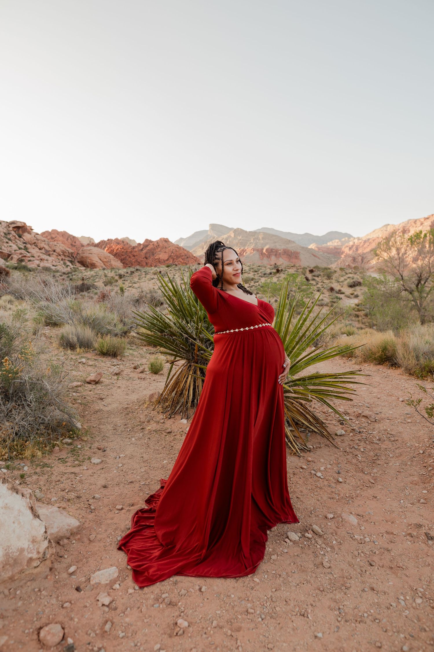 Pregnant woman in a flowing red gown posing at Red Rock Canyon desert landscape.