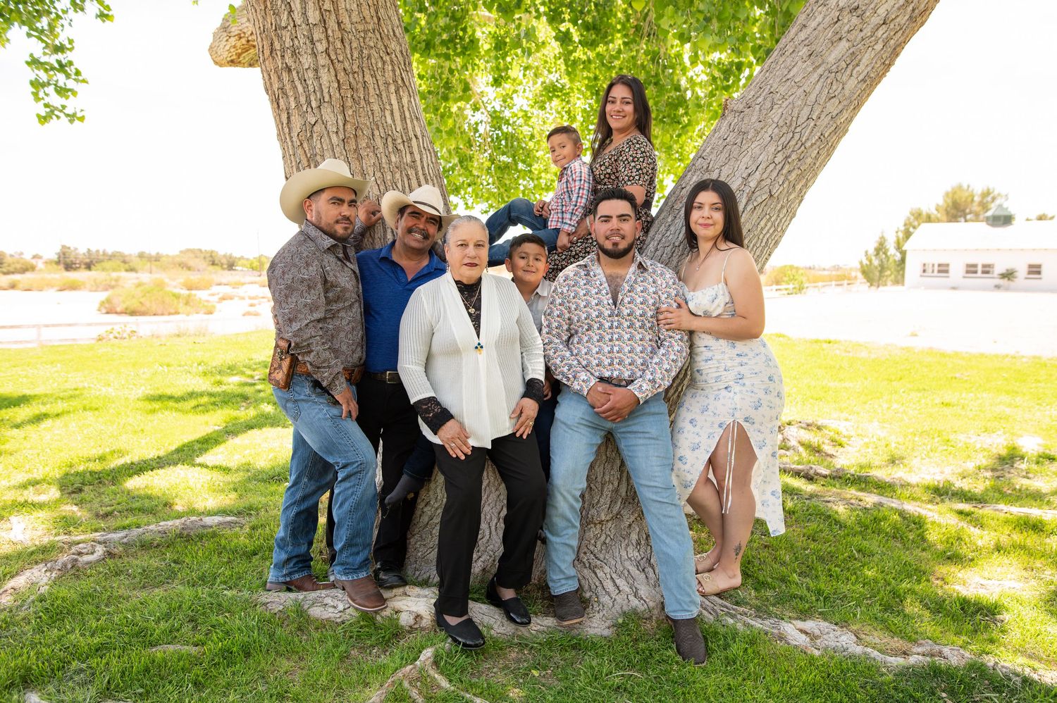 A large family group of eight people poses together in front of a large tree in a sunny park.