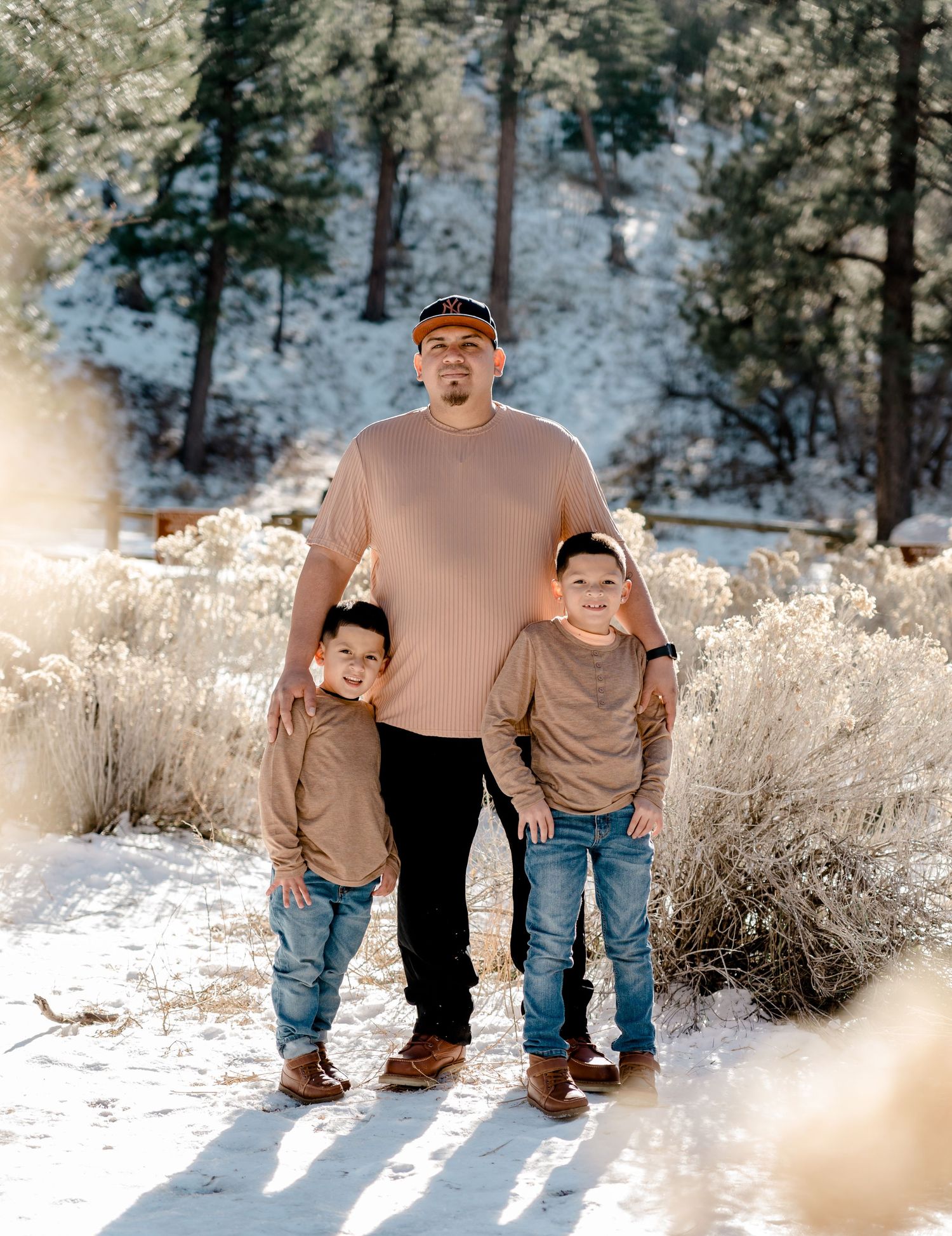Father and two young sons in matching tan shirts posing in snowy pine forest.