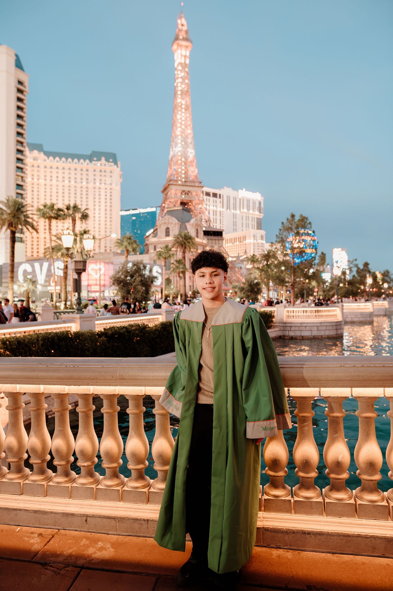 Young man in green graduation gown poses proudly near Paris Las Vegas tower at evening.