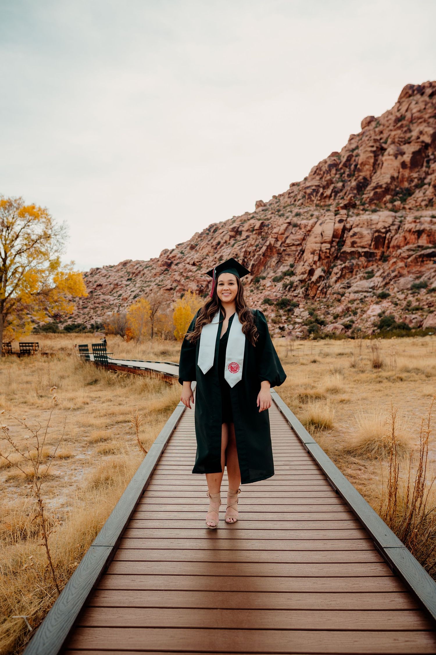 Female graduate in black cap and gown walks wooden boardwalk at Red Rock Canyon Nevada.