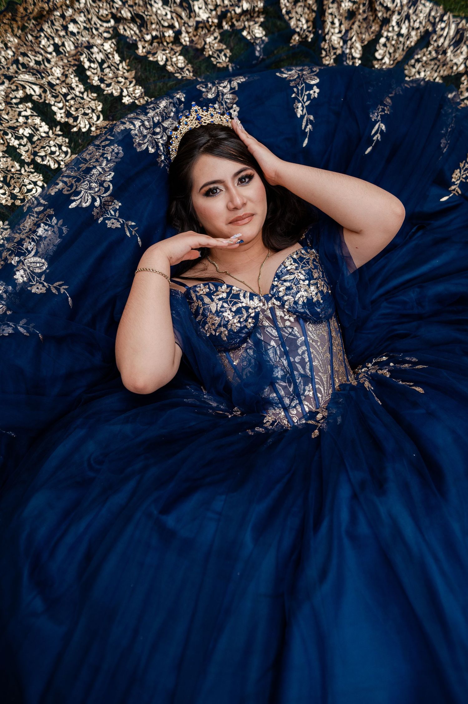 Young woman in navy blue quinceañera ball gown with gold embroidery, lying elegantly among floral decorations.