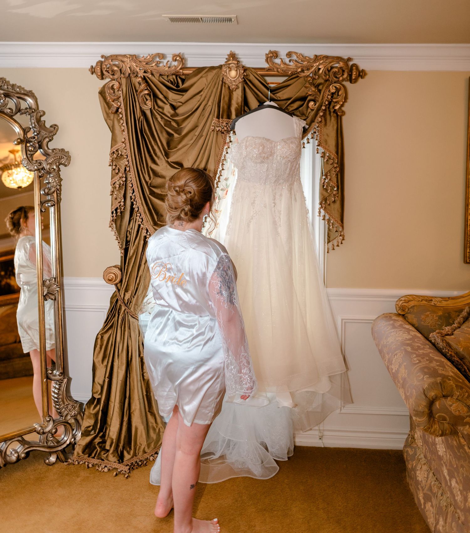 Bride in satin robe admiring her wedding dress hanging on ornate gold curtain rod in elegant dressing room.