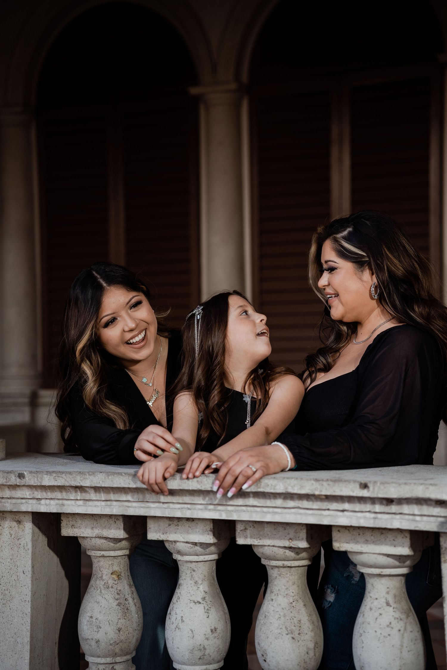 Three females laugh and share a joyful moment together leaning on a white stone balustrade.