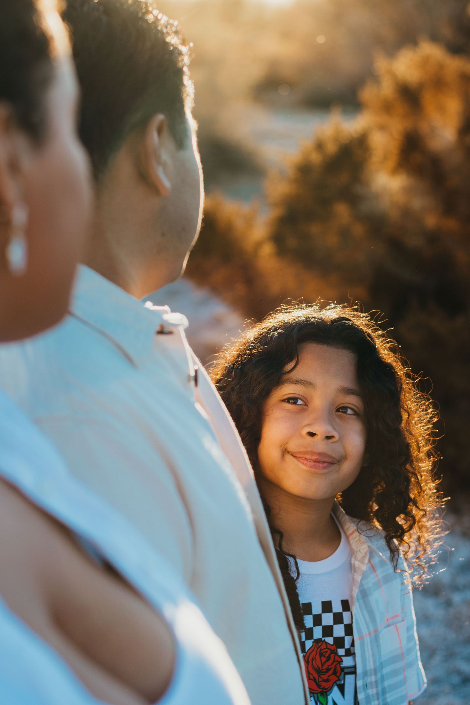 Young child with curly hair smiles softly in warm golden backlit outdoor family portrait.