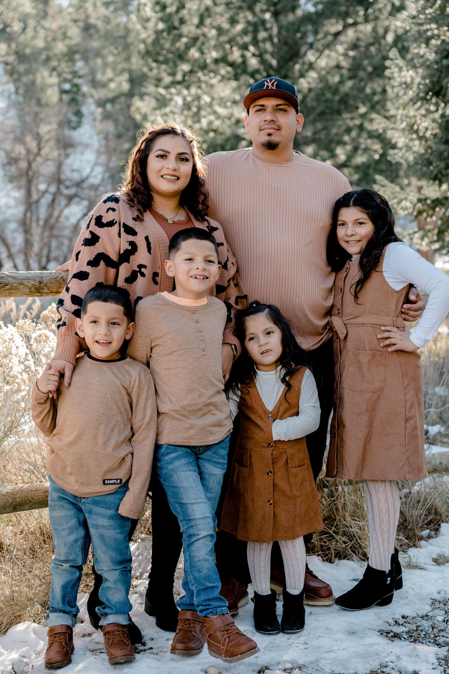 A family of six poses together in a snowy winter forest setting, wearing coordinated earth-tone outfits.