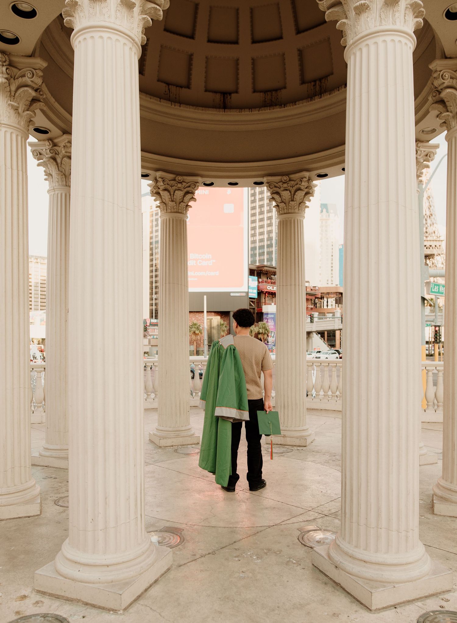 Graduate in green gown standing between ornate white columns at Caesars Palace rotunda.
