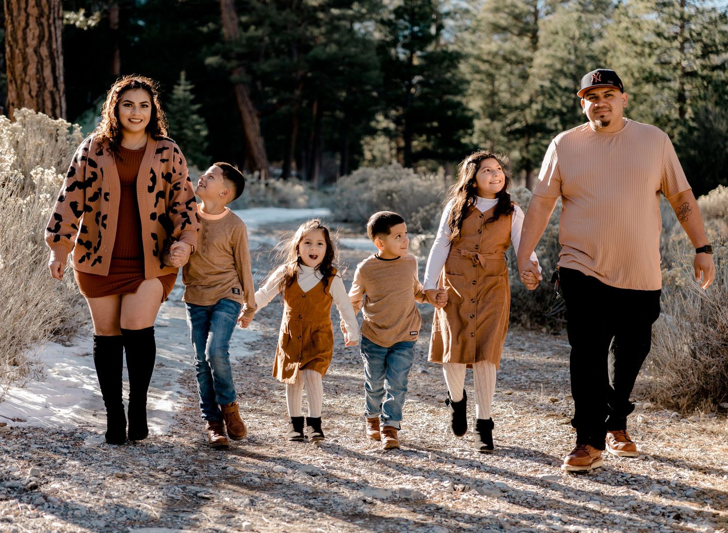 A family of six walks hand-in-hand along a snowy forest path in matching warm-toned autumn outfits.