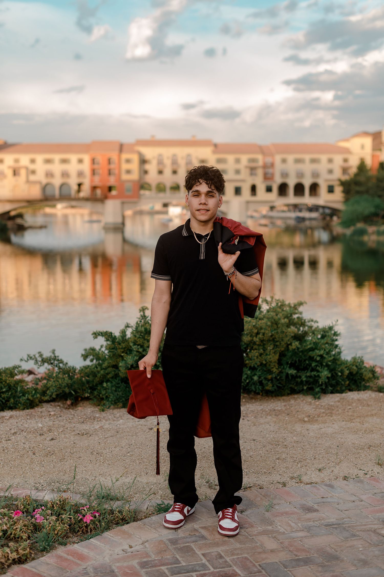 Young man in black outfit holding red graduation gown by lakeside at sunset.