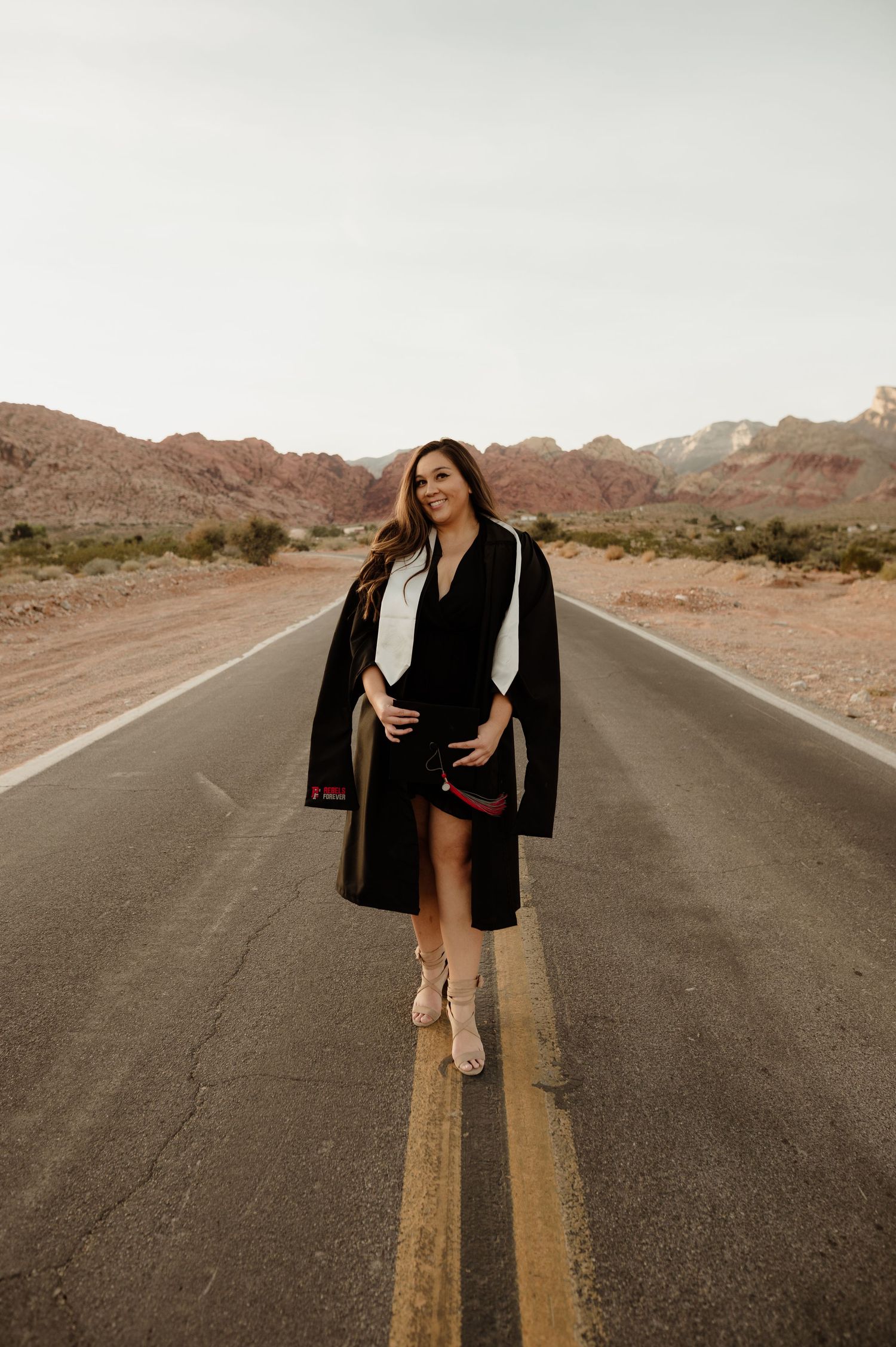 Woman in graduation gown walks down empty desert road with red rock mountains in background.