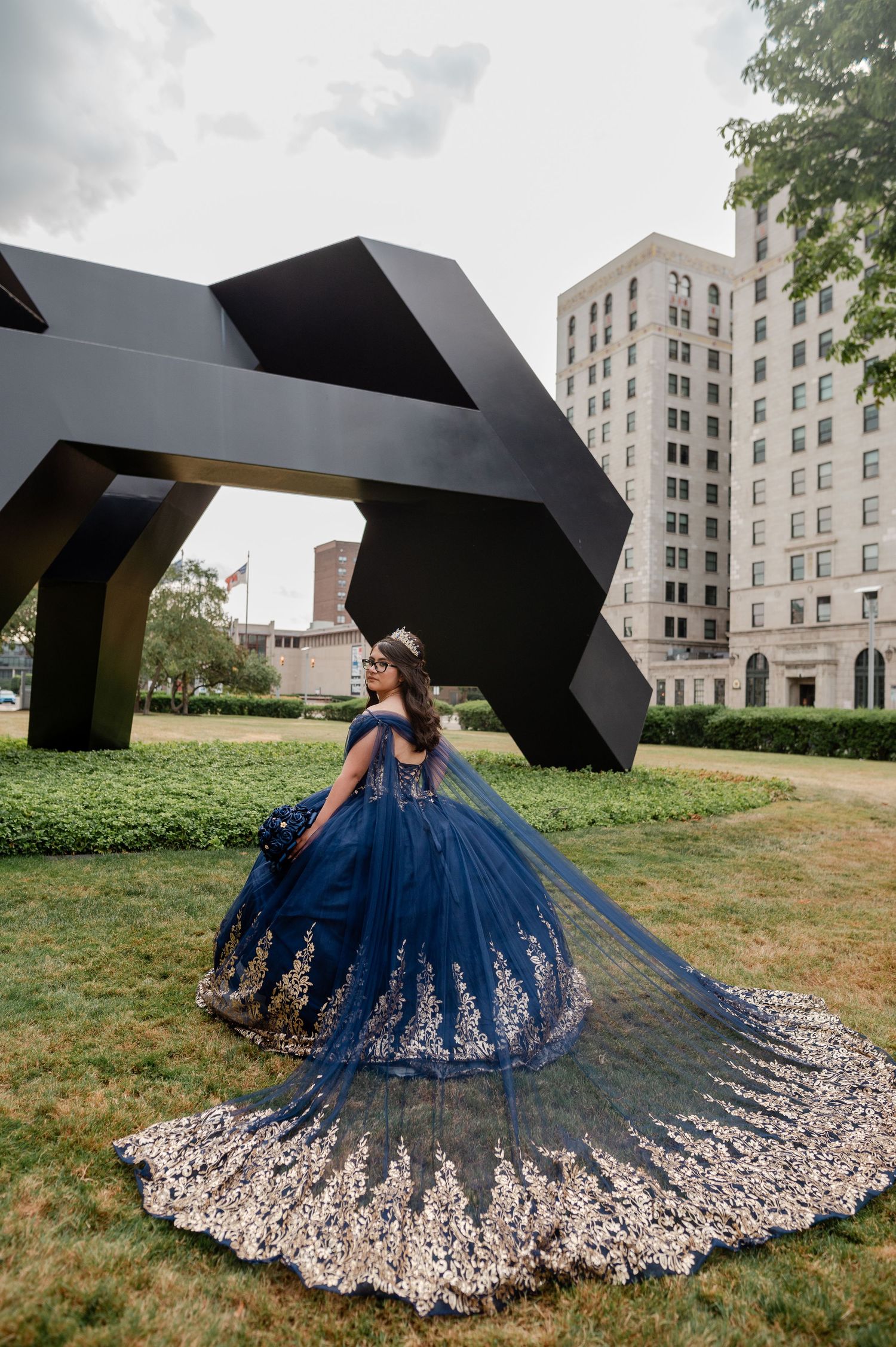 Young woman in navy blue quinceañera gown with gold lace trim poses outdoors near modern sculpture and city building.