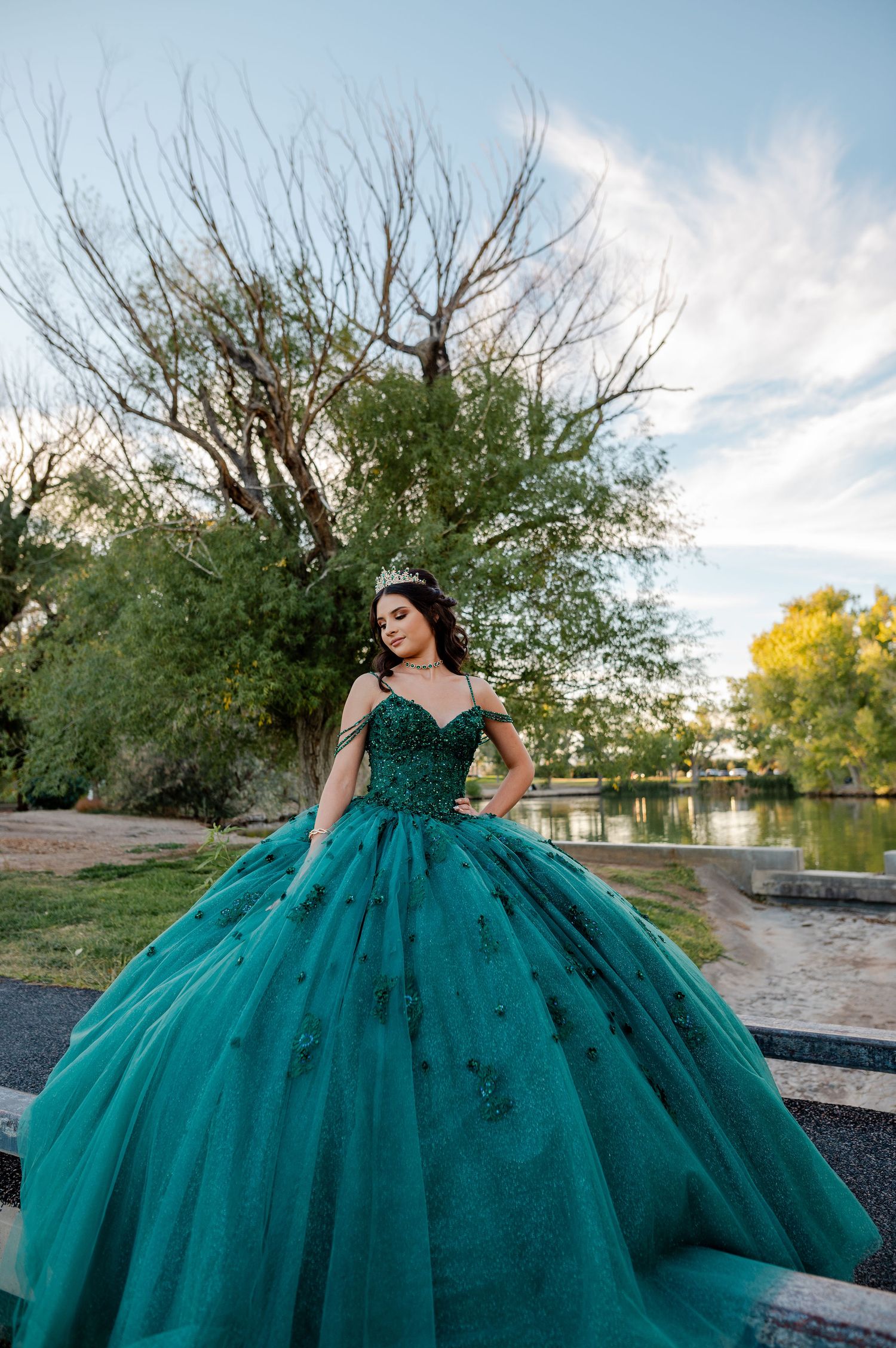 Young woman in teal quinceañera ball gown with floral appliqués poses near a lakeside park.