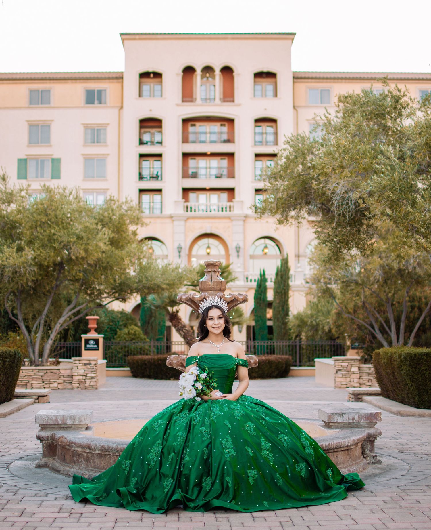 Young woman in an emerald green quinceañera ball gown poses in front of an elegant hotel.