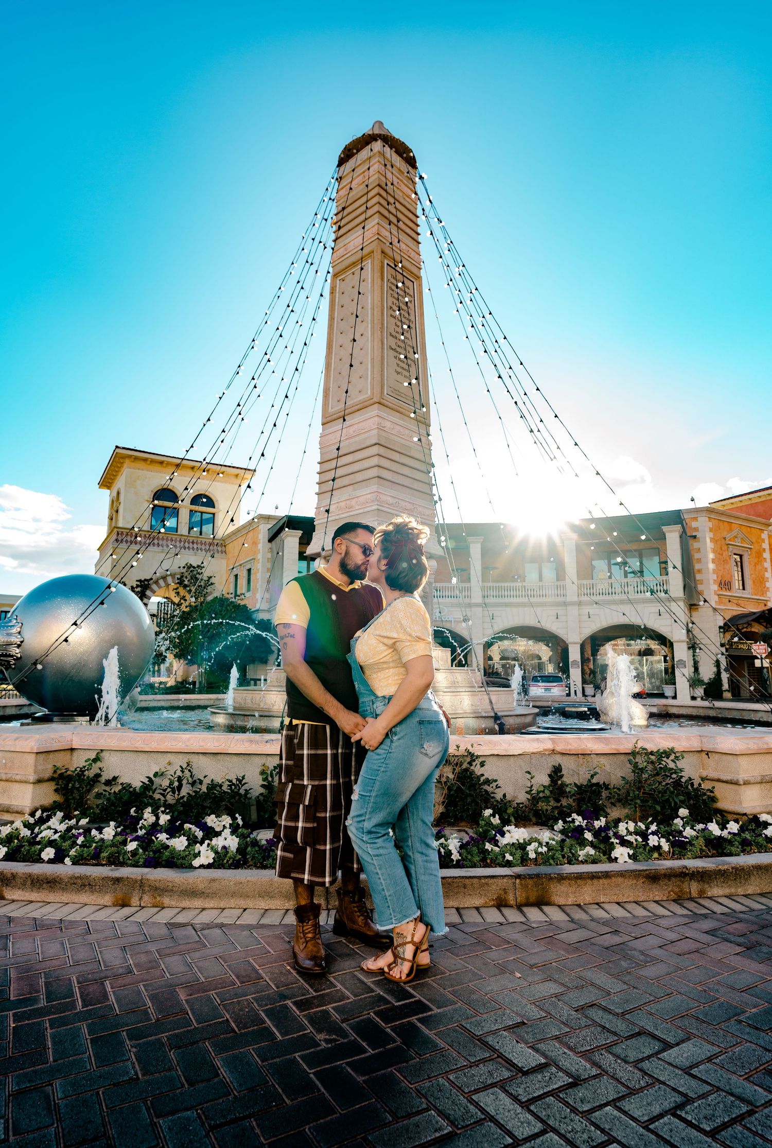 Couple kissing near a fountain with a tall tower and sunburst in a plaza.