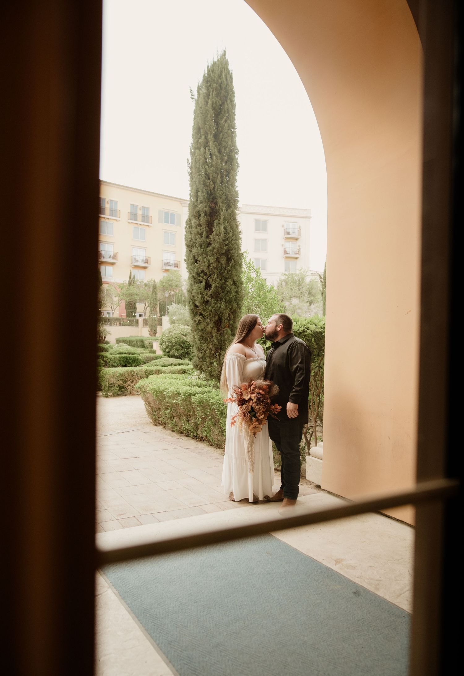 Bride and groom share a romantic moment framed by an arched doorway with lush garden backdrop.