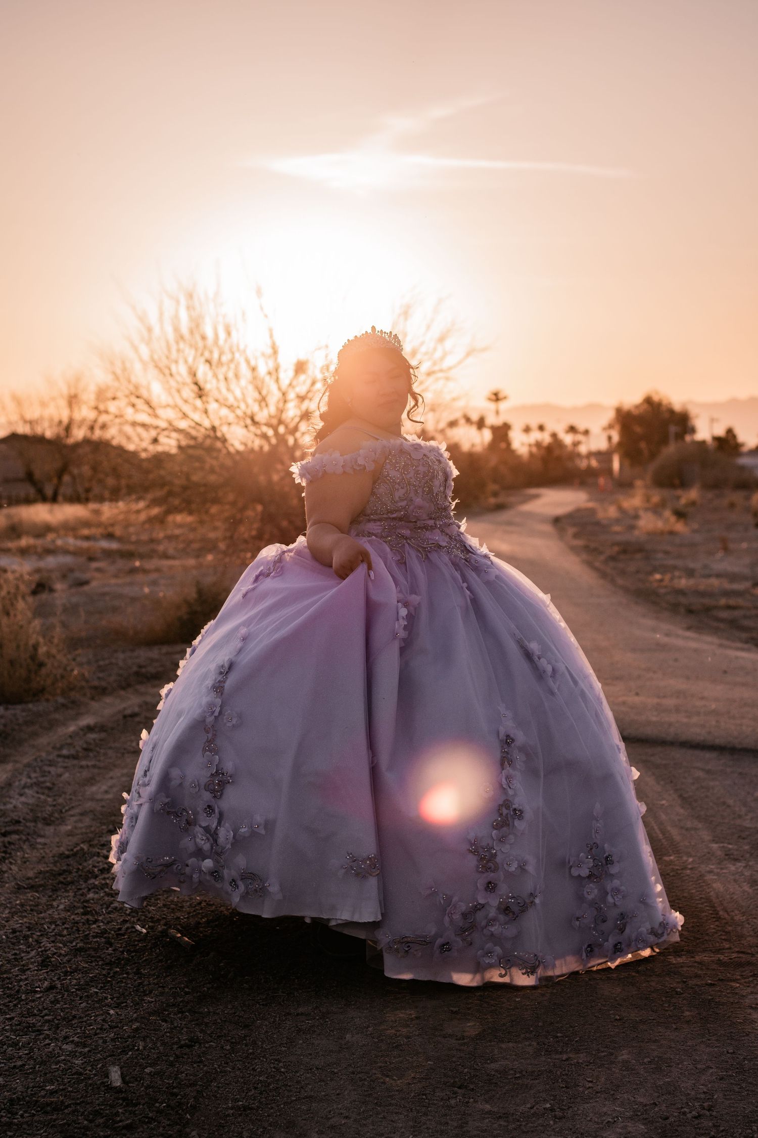 Quinceañera in lavender floral ball gown walks desert path backlit by golden sunset glow.
