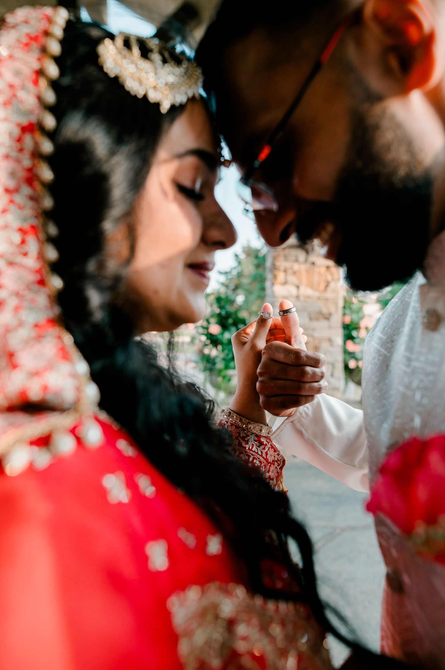 Indian bride in red lehenga and groom in white share an intimate moment holding hands during traditional wedding ceremony.