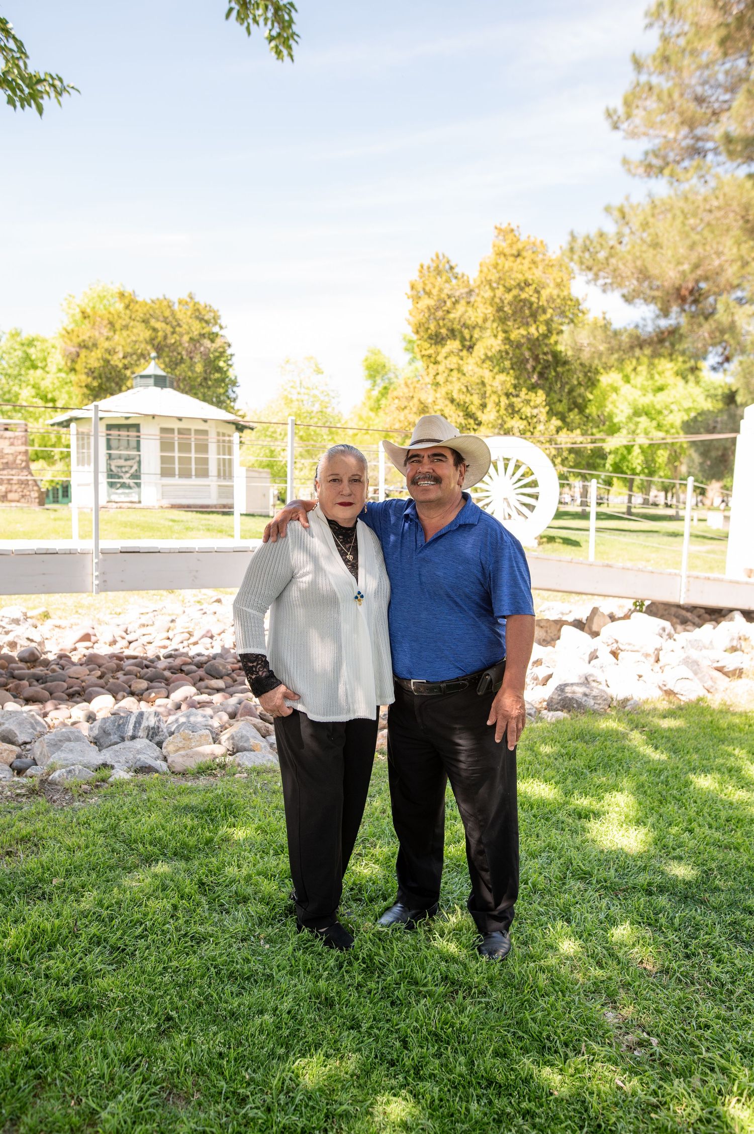An older couple smiles together in a green park setting with a gazebo and wagon wheel visible behind them.