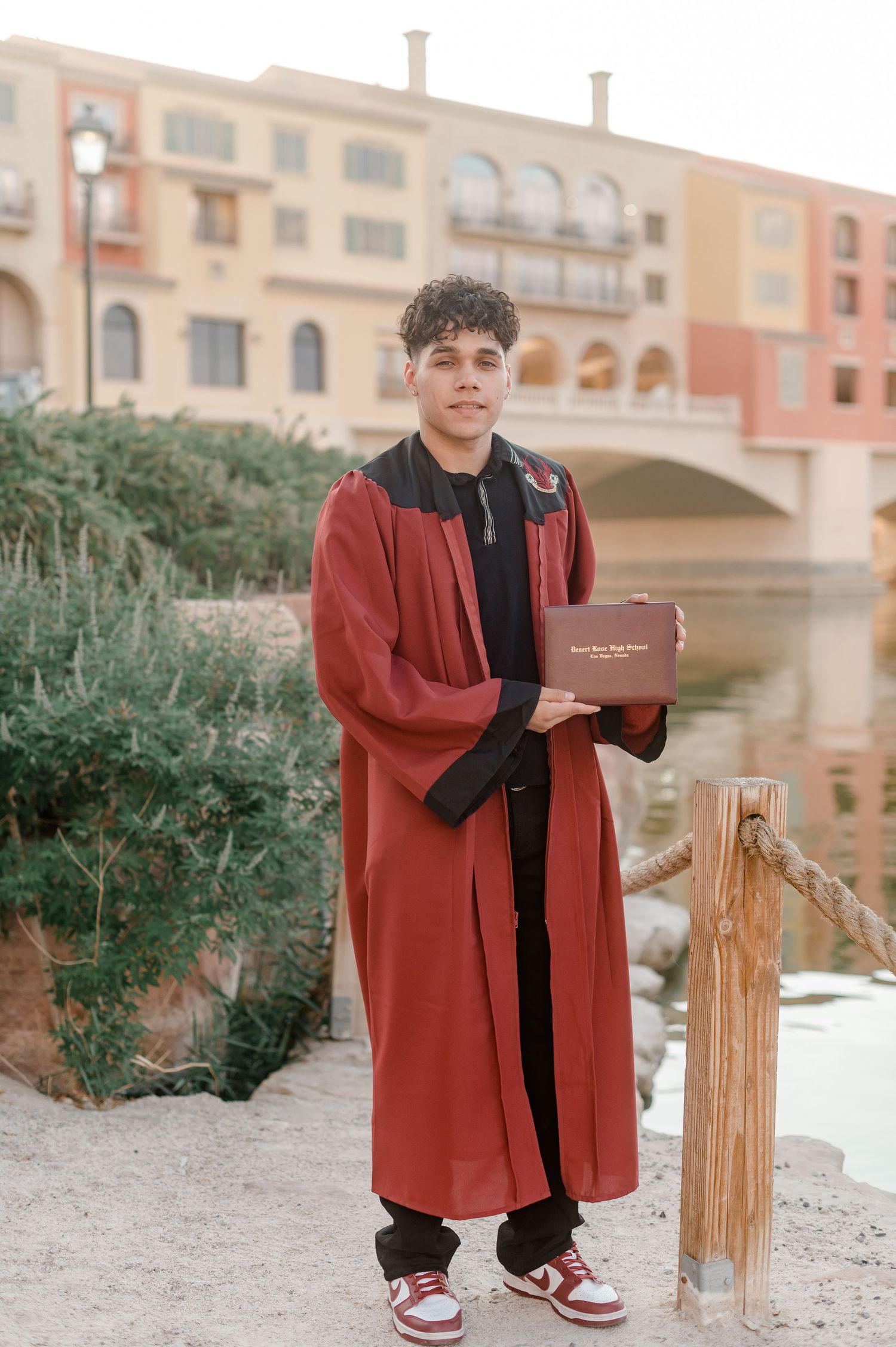 Proud graduate in red gown holds diploma cover by waterfront with colorful buildings behind.