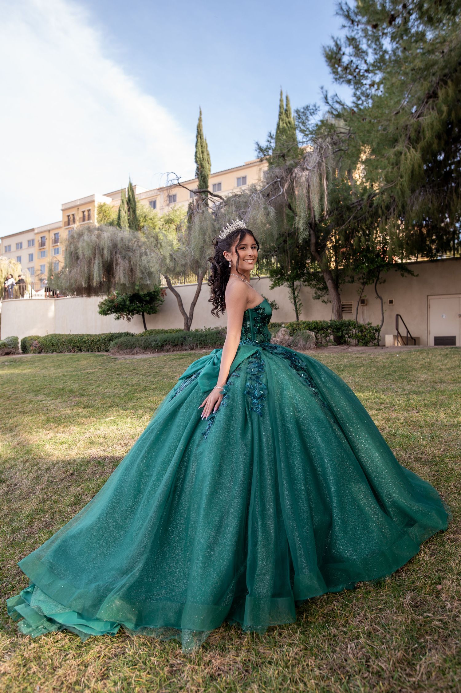 Beautiful quinceañera twirling in emerald green ball gown with crown in sunny outdoor garden setting.