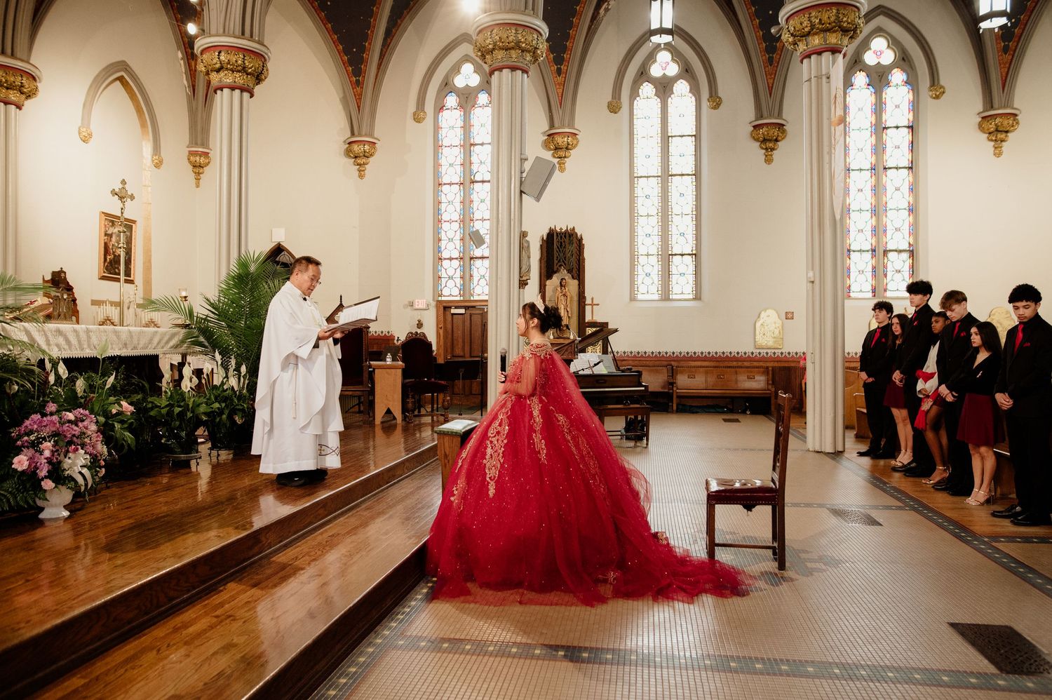 Quinceañera girl in red gown stands before priest at church ceremony.