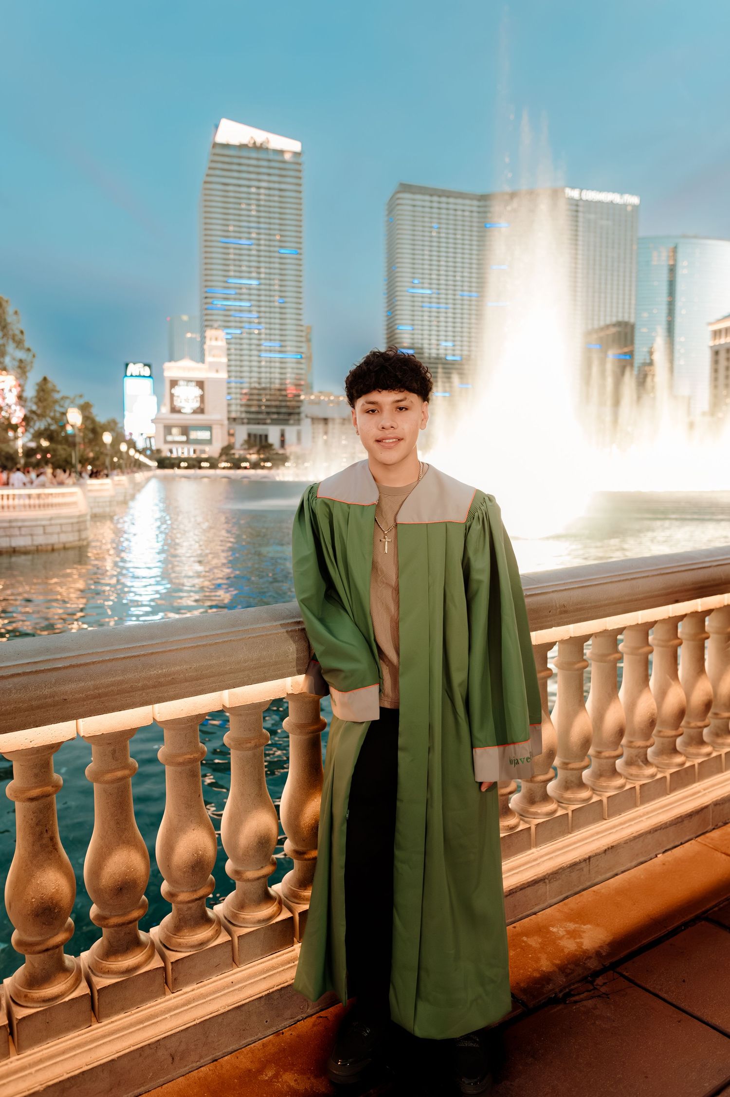 Young man in green graduation gown poses by Bellagio fountain on Las Vegas Strip at dusk.