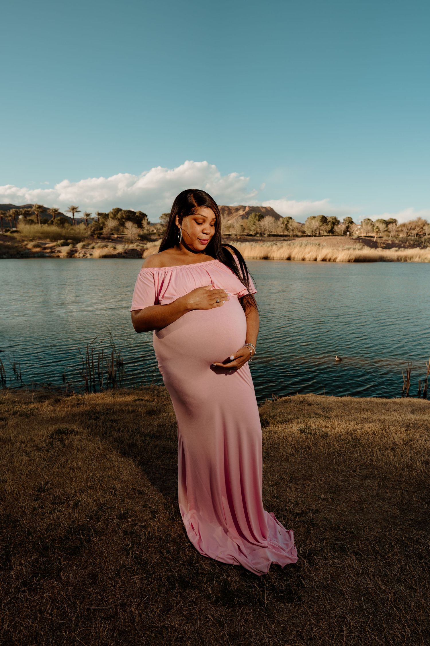 Expectant mother in elegant pink maternity dress holding her belly near a serene lake at golden hour.