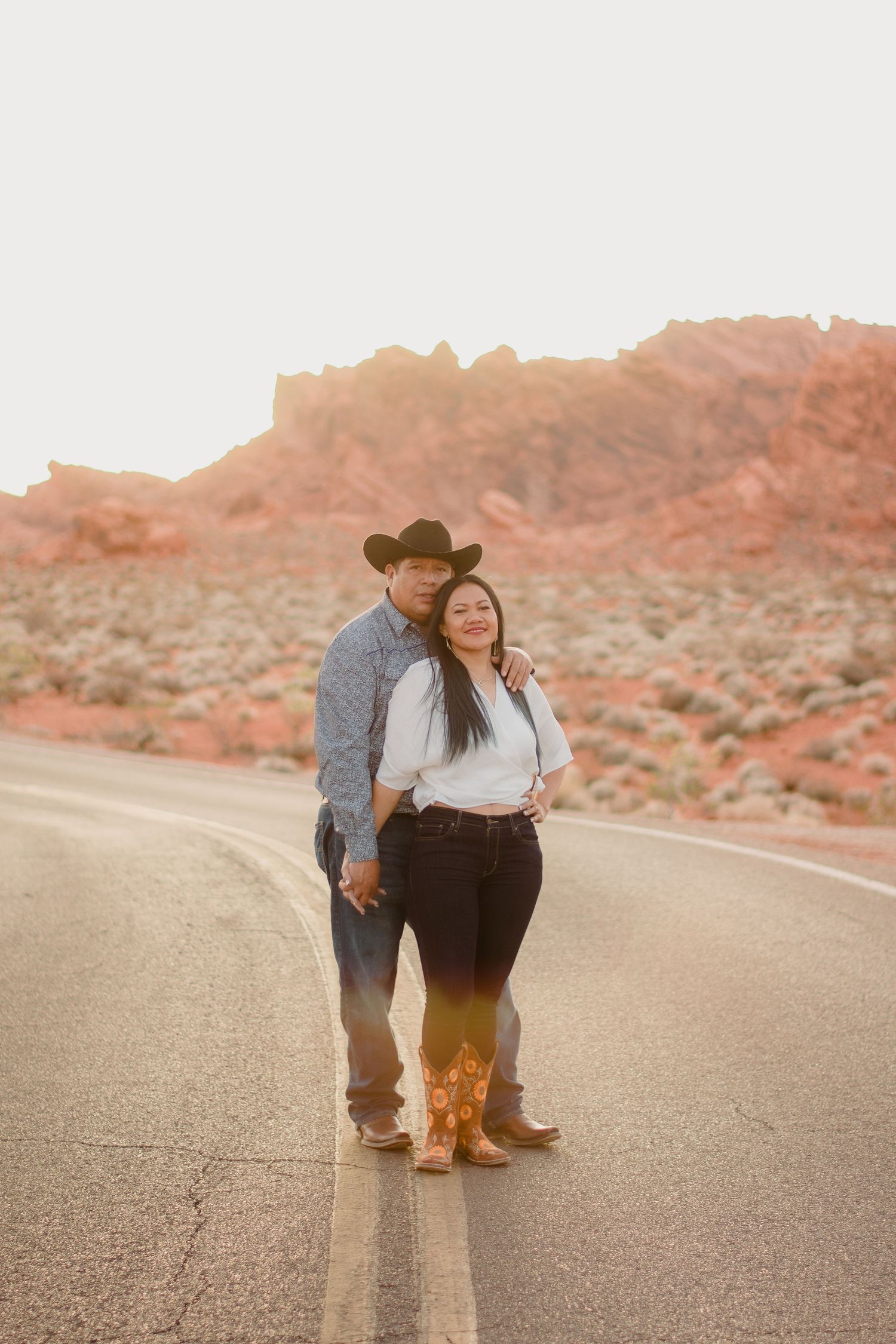 Couple in cowboy hats posing on desert road at sunset with red rock landscape backdrop.