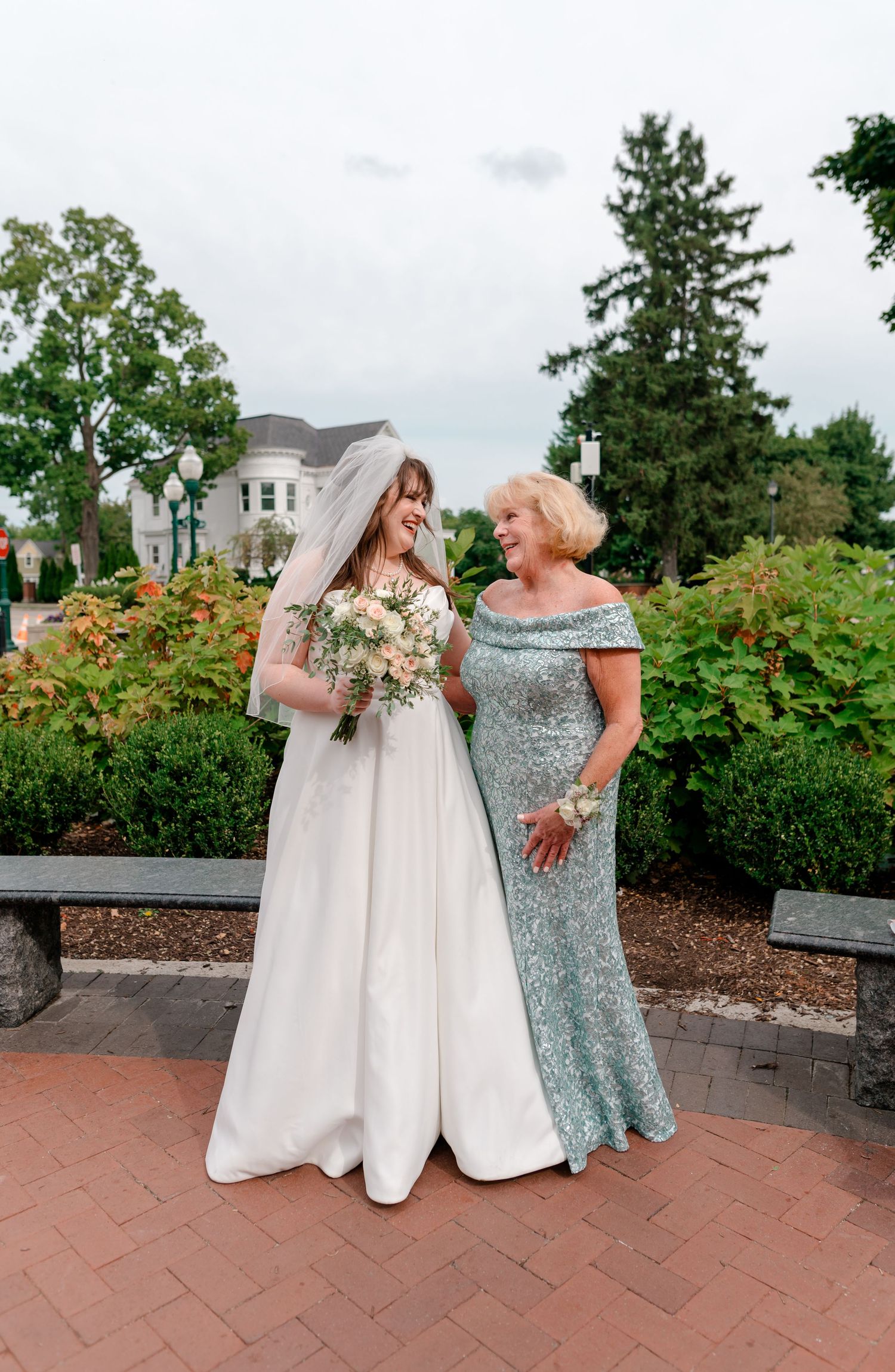 Bride in white gown and veil laughing with mother in teal sequin dress holding bouquet in garden setting.