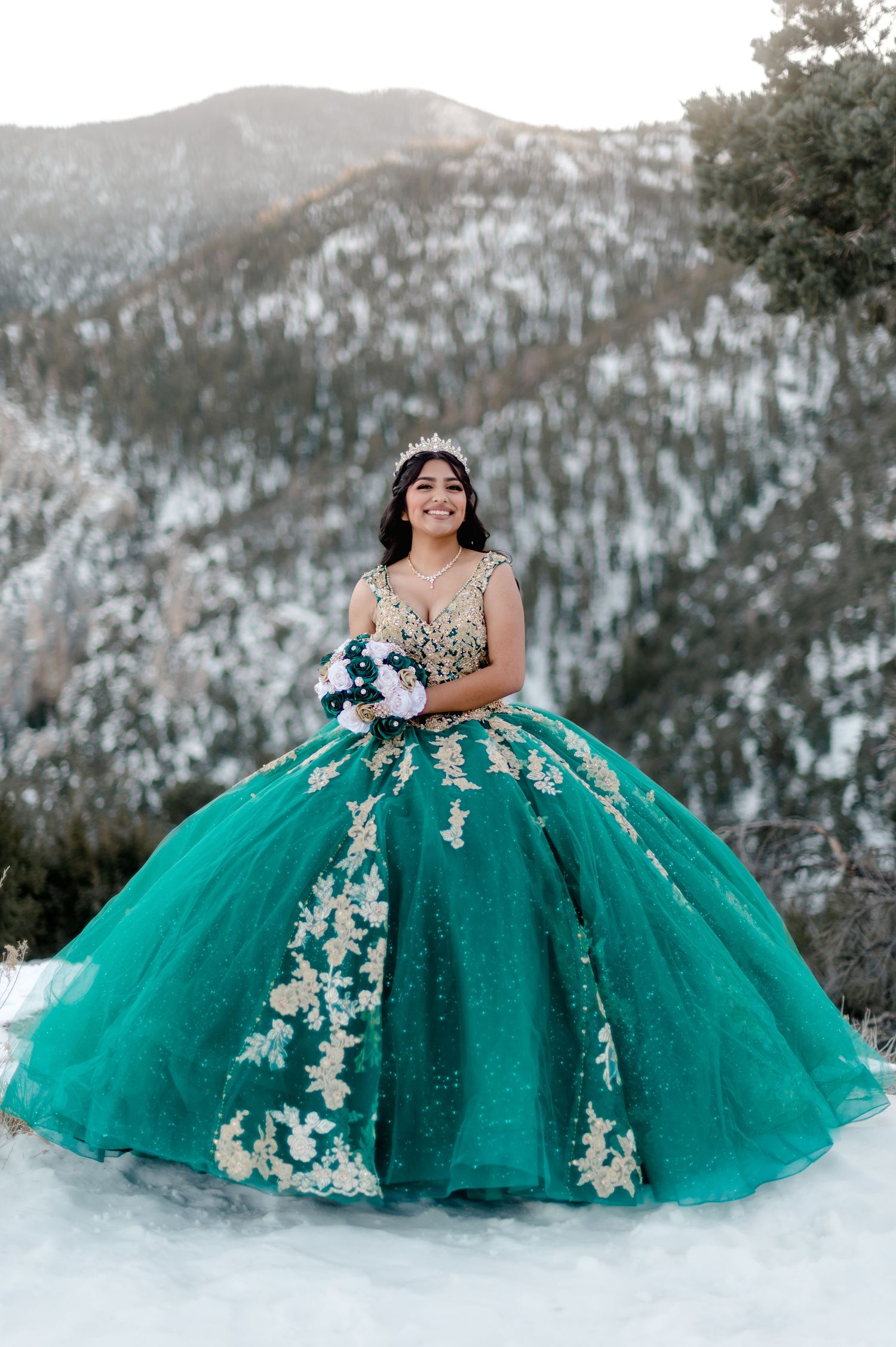Young woman in emerald green quinceañera gown with gold lace holds bouquet on a snowy mountain.