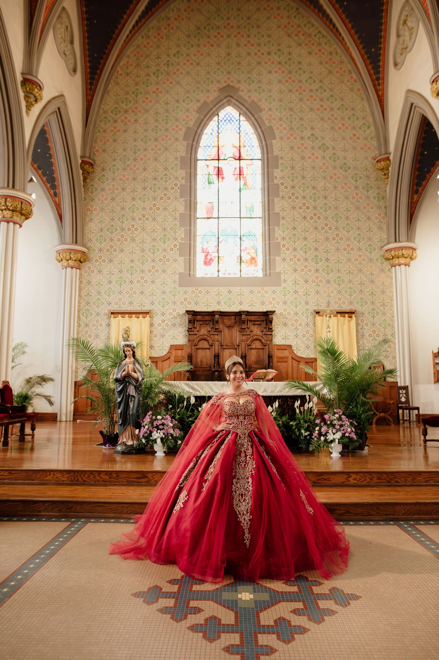 Quinceañera in red and gold ball gown posing at church altar with statue of Virgin Mary nearby.
