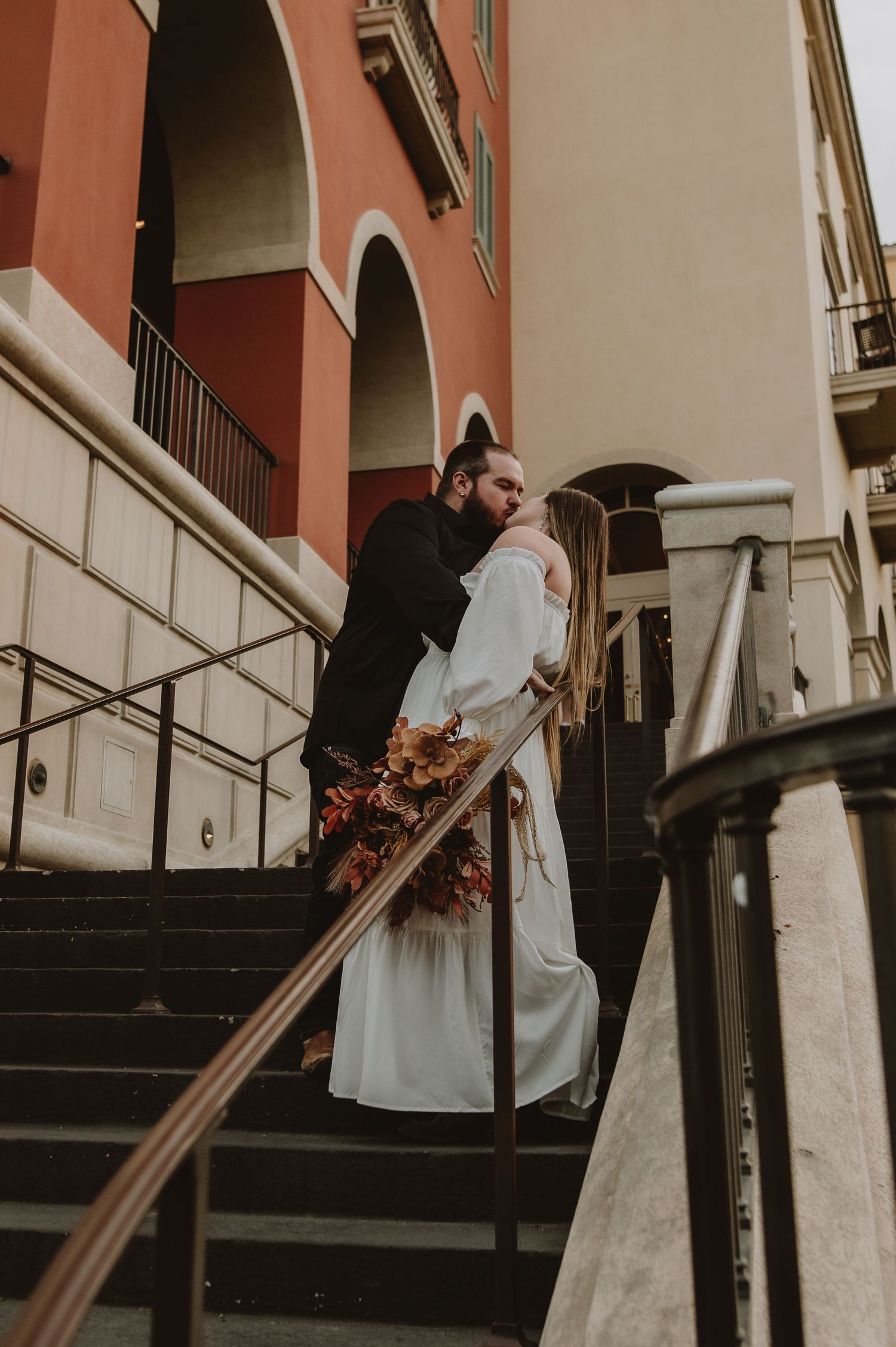Couple shares romantic kiss on outdoor staircase beside a warm terracotta building with ornate railings.