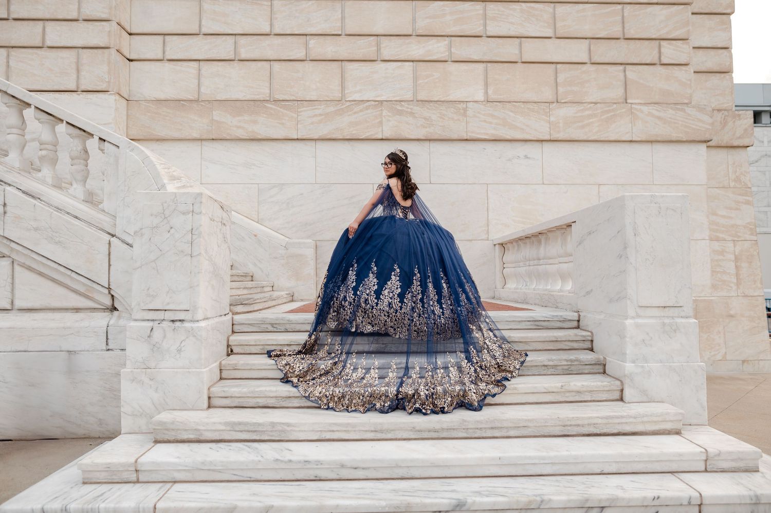 Young woman in navy blue quinceañera ball gown with gold embroidery poses on marble staircase steps.