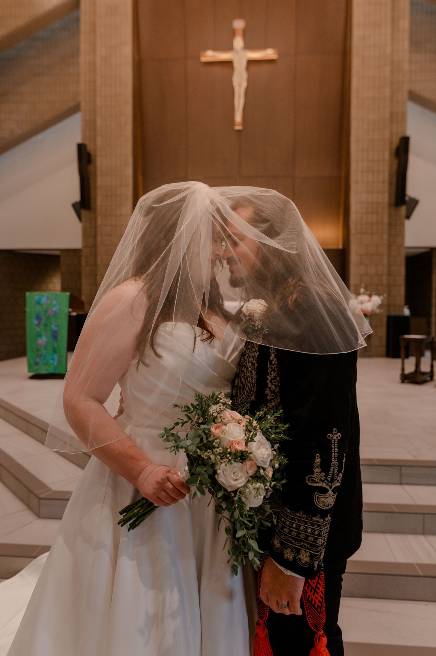 Bride and groom share first kiss under veil at church altar with crucifix, bride holding white floral bouquet.