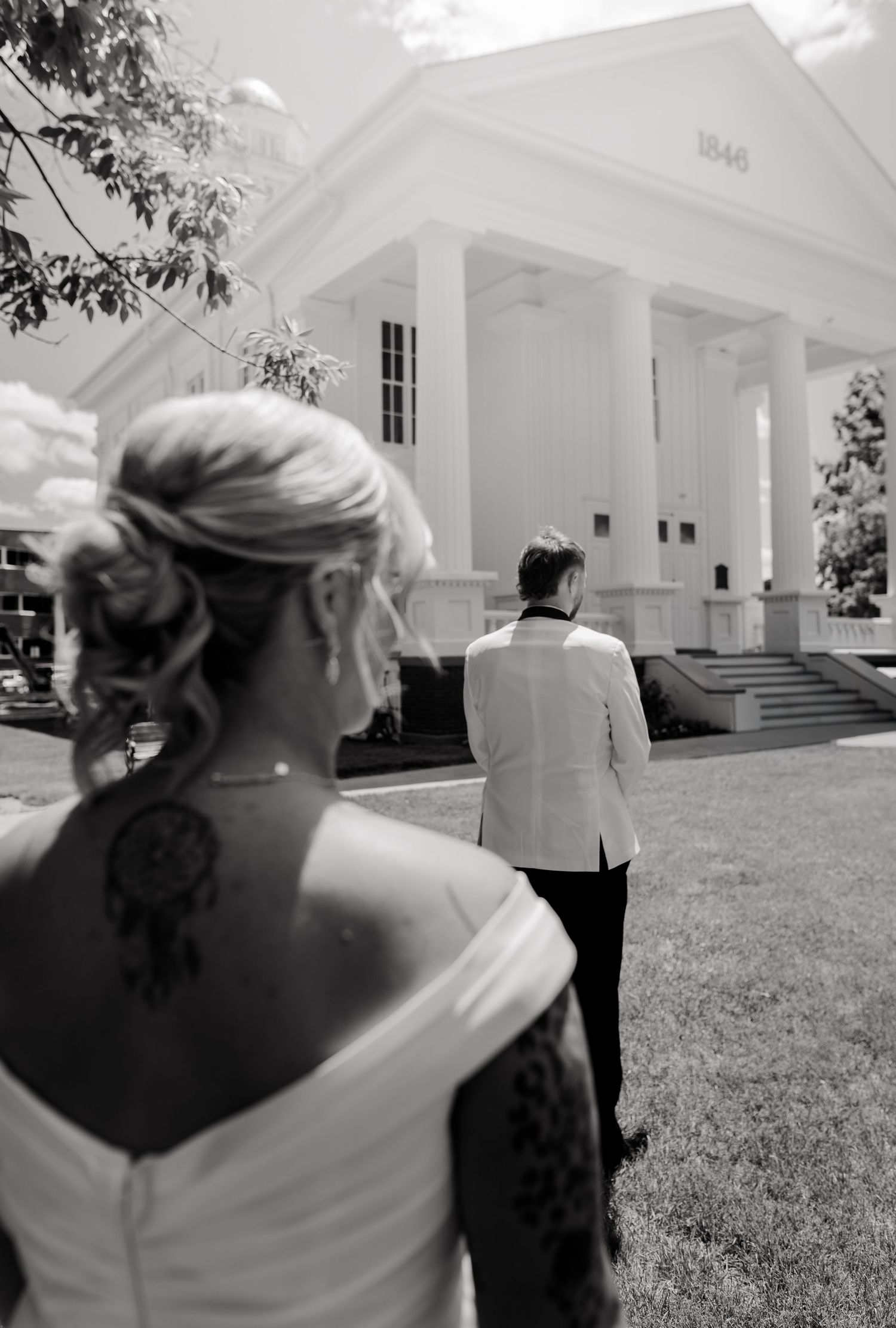 Black and white photo of bride with dream catcher tattoo facing groom in white tuxedo outside historic white columned church.