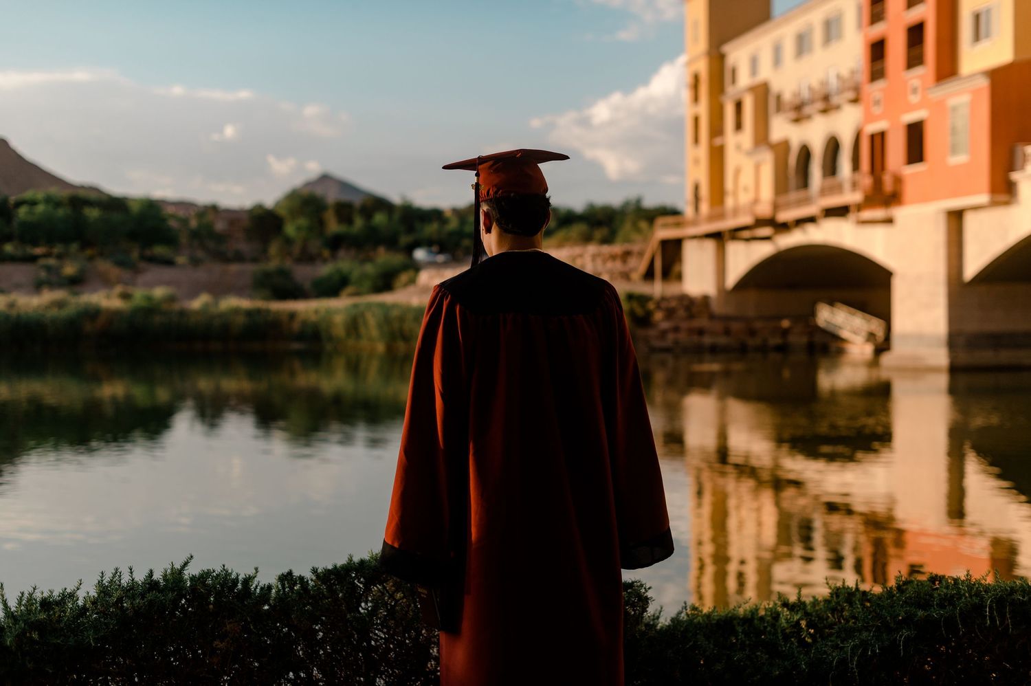 Graduate in red cap and gown stands contemplatively overlooking a serene lake at golden hour.