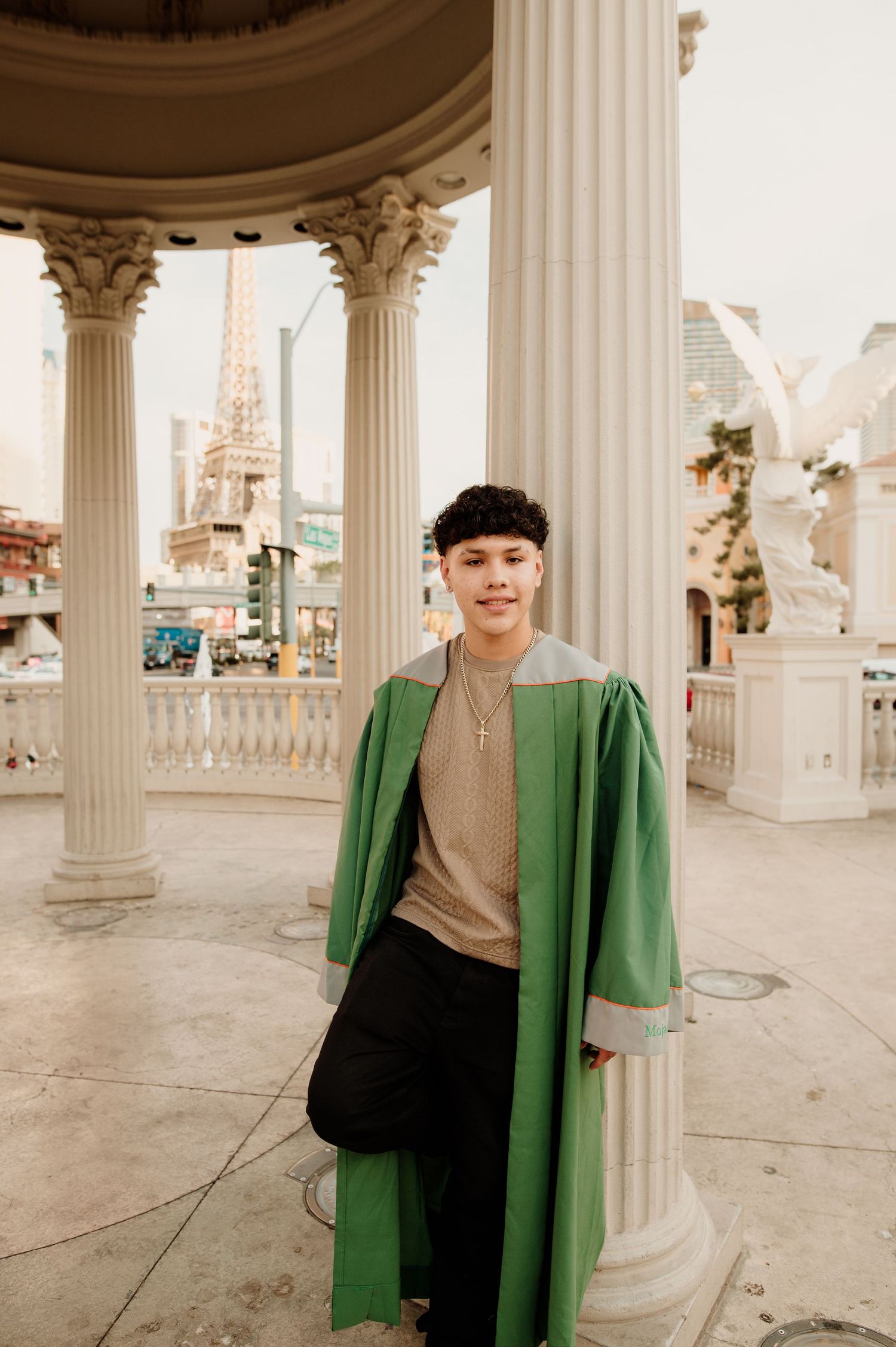 Smiling graduate in green gown standing among classical columns with Eiffel Tower replica behind.