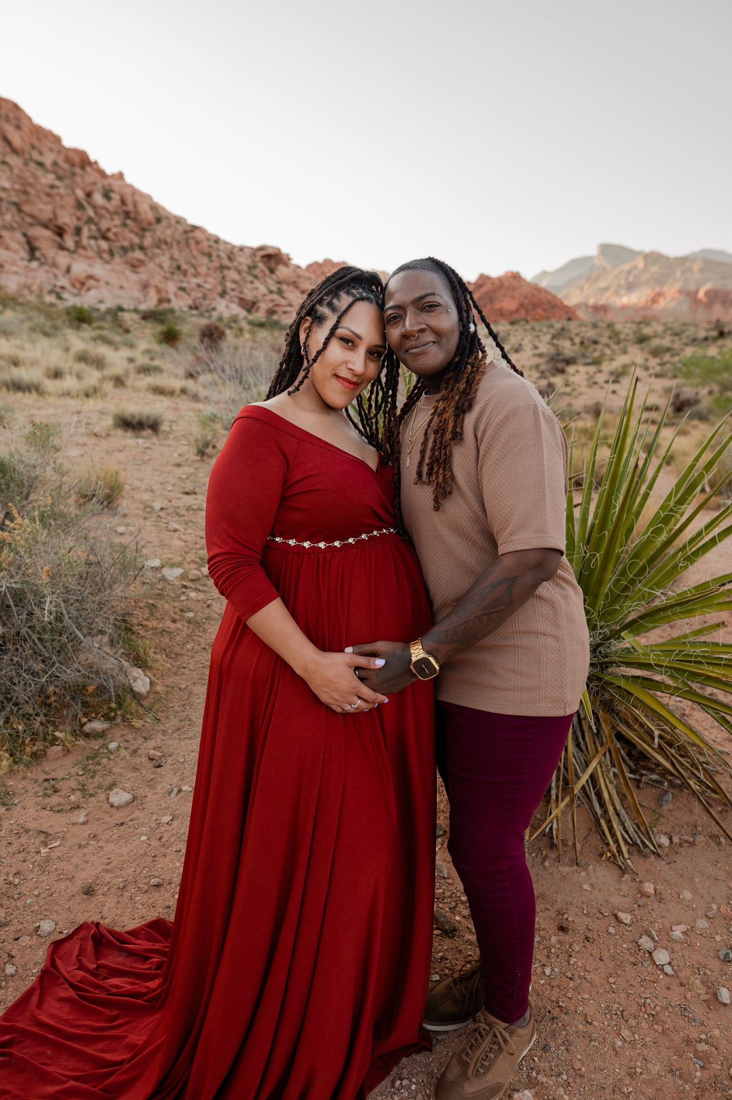 Couple poses together in Red Rock Canyon desert, woman wearing elegant red maternity gown.