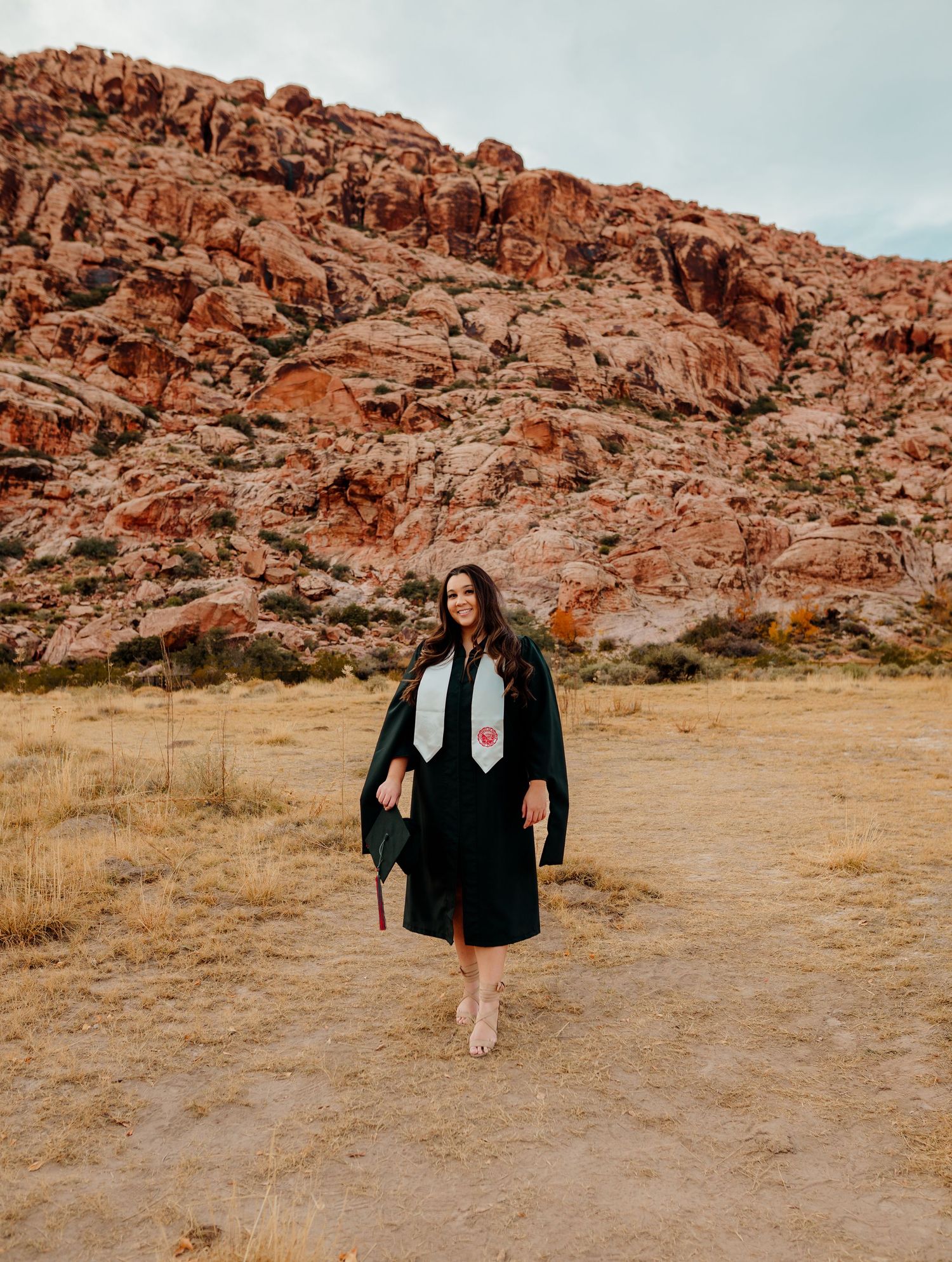 Woman in black graduation gown and stole walks through Red Rock Canyon desert landscape.