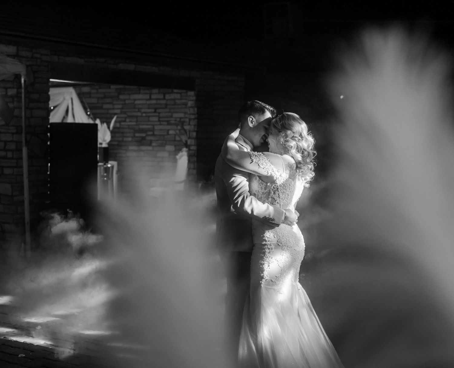 Bride and groom sharing romantic first dance embrace surrounded by atmospheric fog in black and white.