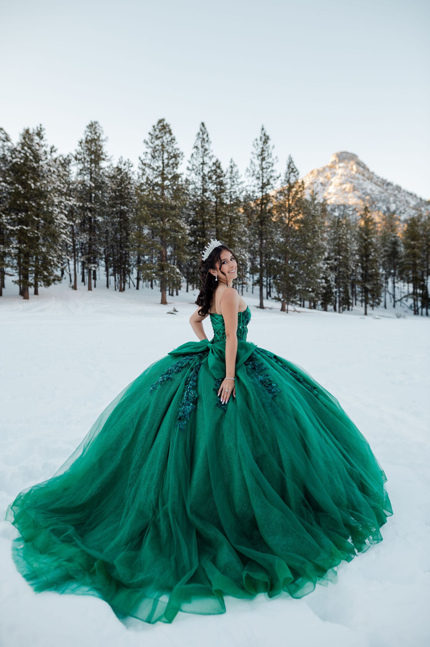 Young woman in voluminous strapless emerald green quinceañera gown posing in snowy mountain setting.