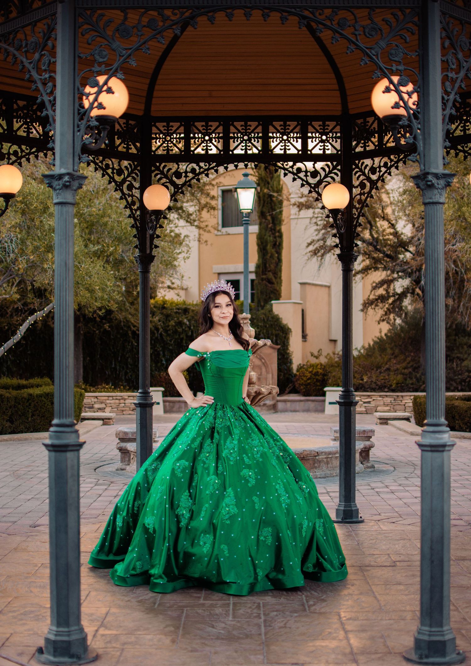 Young woman in a green quinceañera ball gown poses under an ornate iron gazebo at dusk.